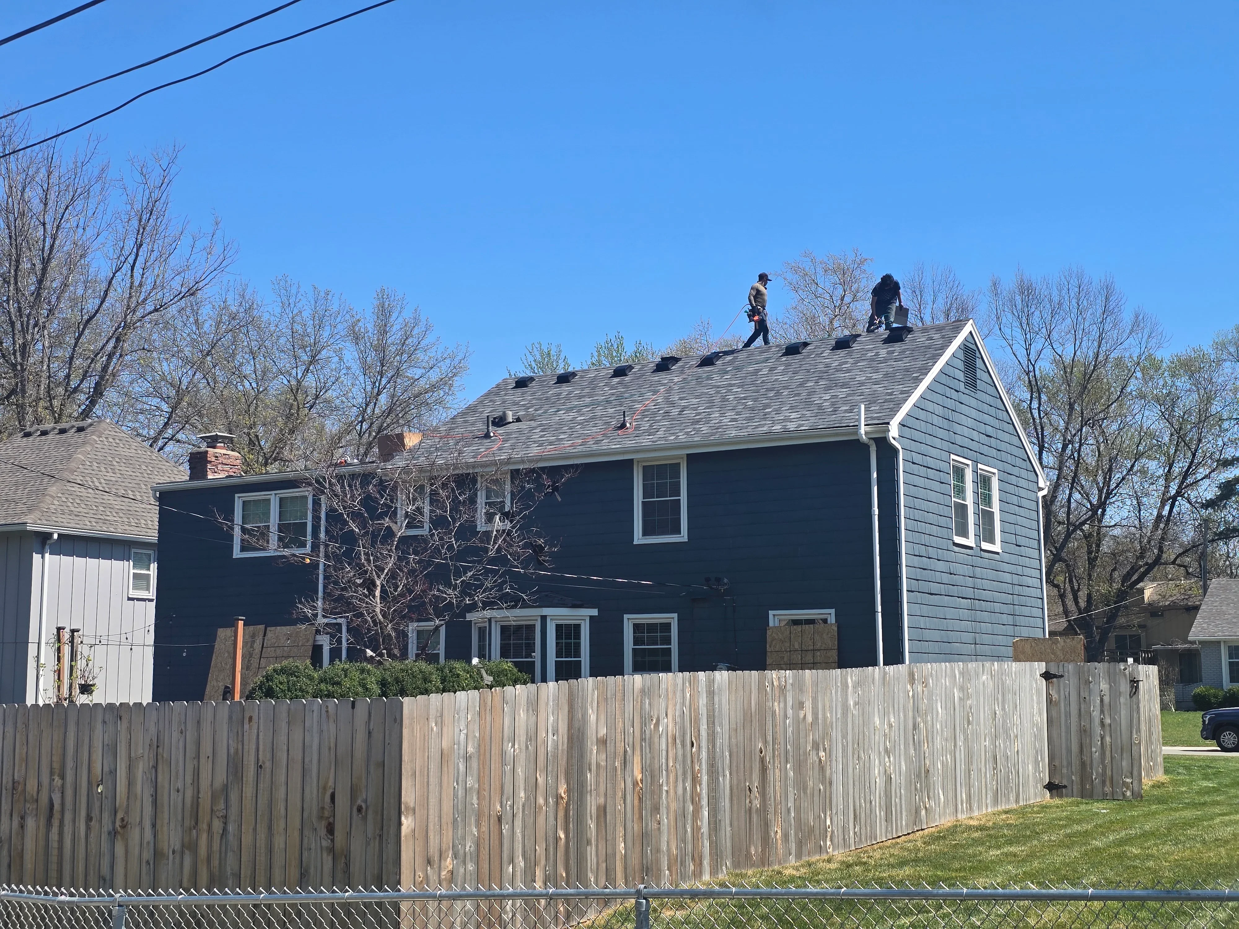 Two workers installing gray asphalt shingles on residential roof with white trim and dark blue siding